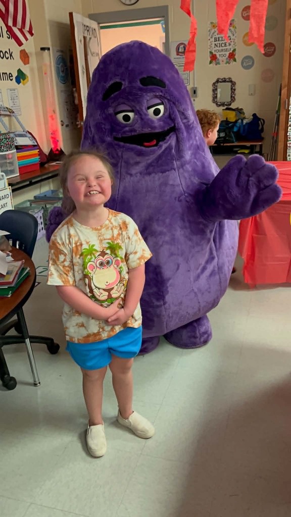 A child stands next to a large purple mascot with a smiling face in a classroom.