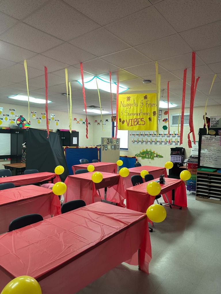 A classroom with tables covered in red cloth. Each table has yellow balloons attached to them.