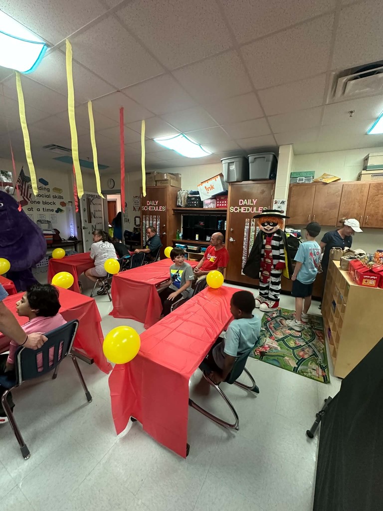 A classroom with tables covered in red cloth, balloons, and several children seated. Ceiling features hanging yellow streamers.