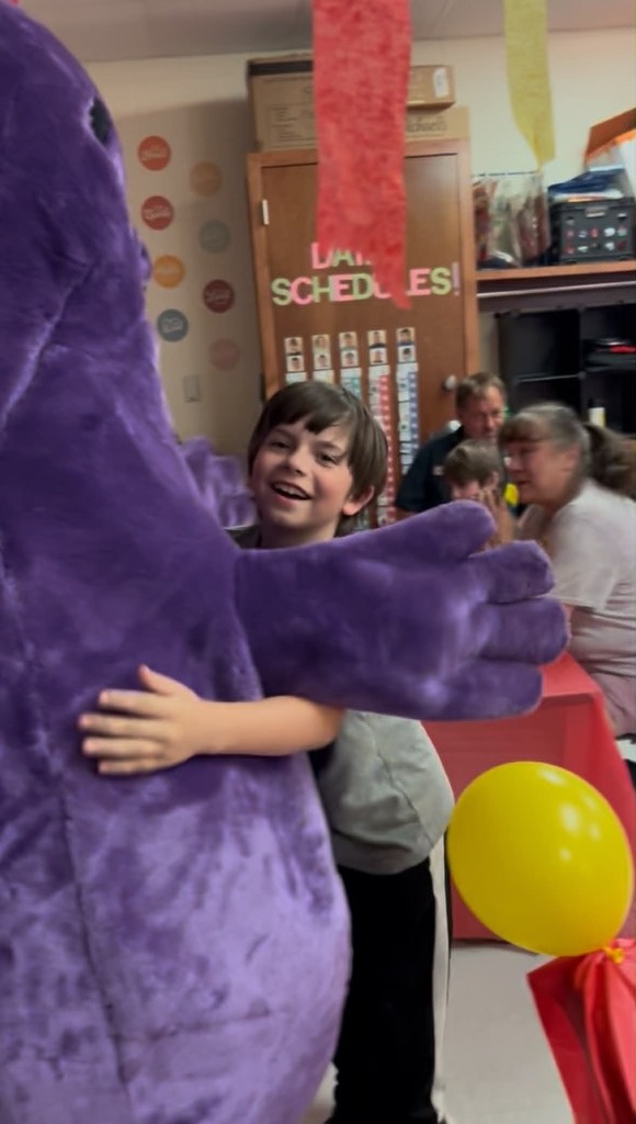 A child in a gray shirt hugs a purple mascot. Behind them, adults and children sit around a table with a yellow balloon.