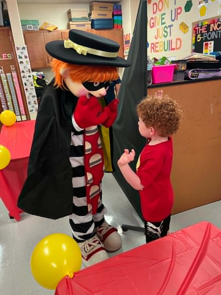 A mascot dressed in a clown costume with a red cape stands next to a young child. They are near a table with red tablecloth and yellow balloons. Behind them is a counter with a pink bucket.