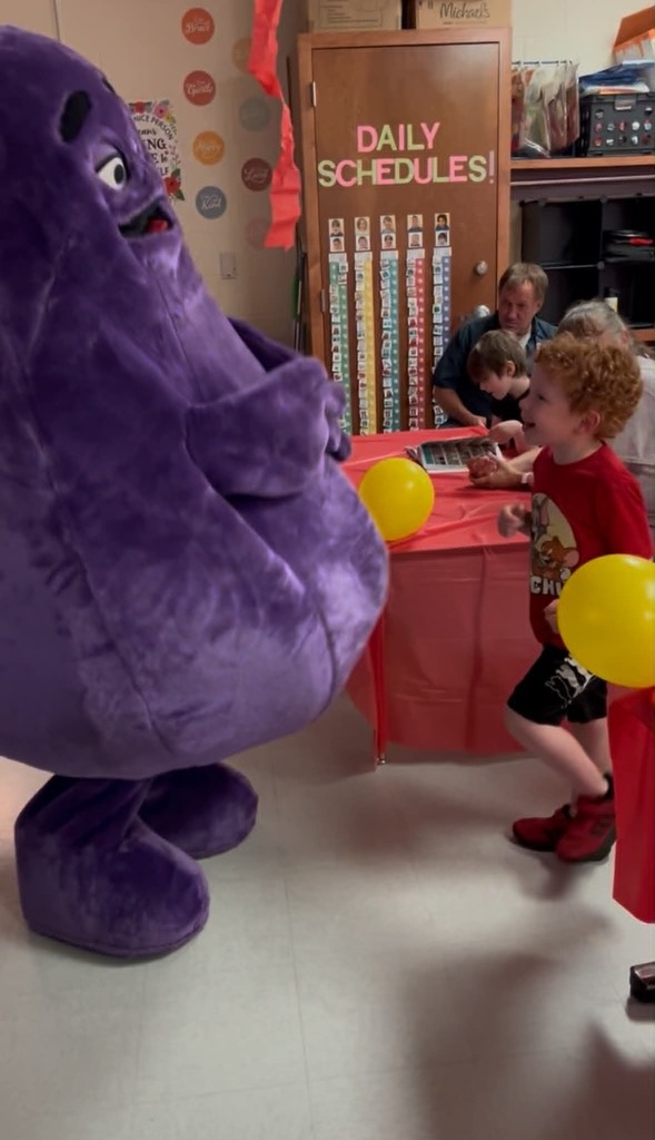 A purple mascot stands near a table, balloons, and a boy. Two adults sit at a table behind the mascot.