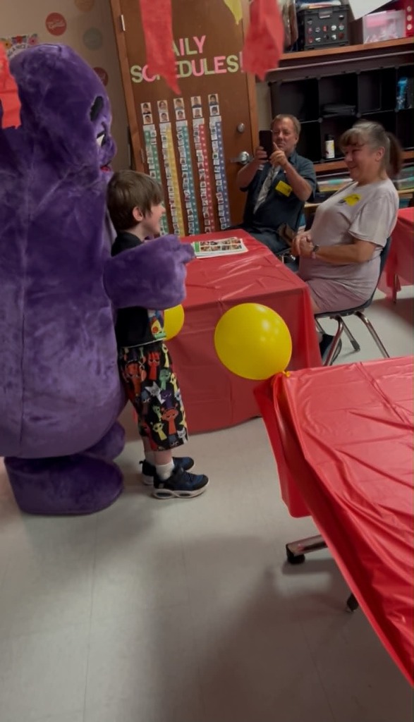 A child stands next to a mascot with a balloon. Two adults sit at a table. The walls display a bulletin board.