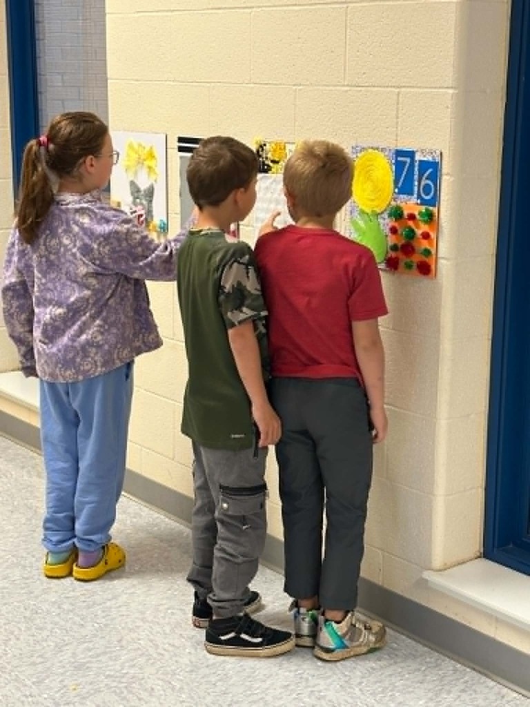Three children examining artwork on a wall. The artwork includes paintings of flowers and numbers.