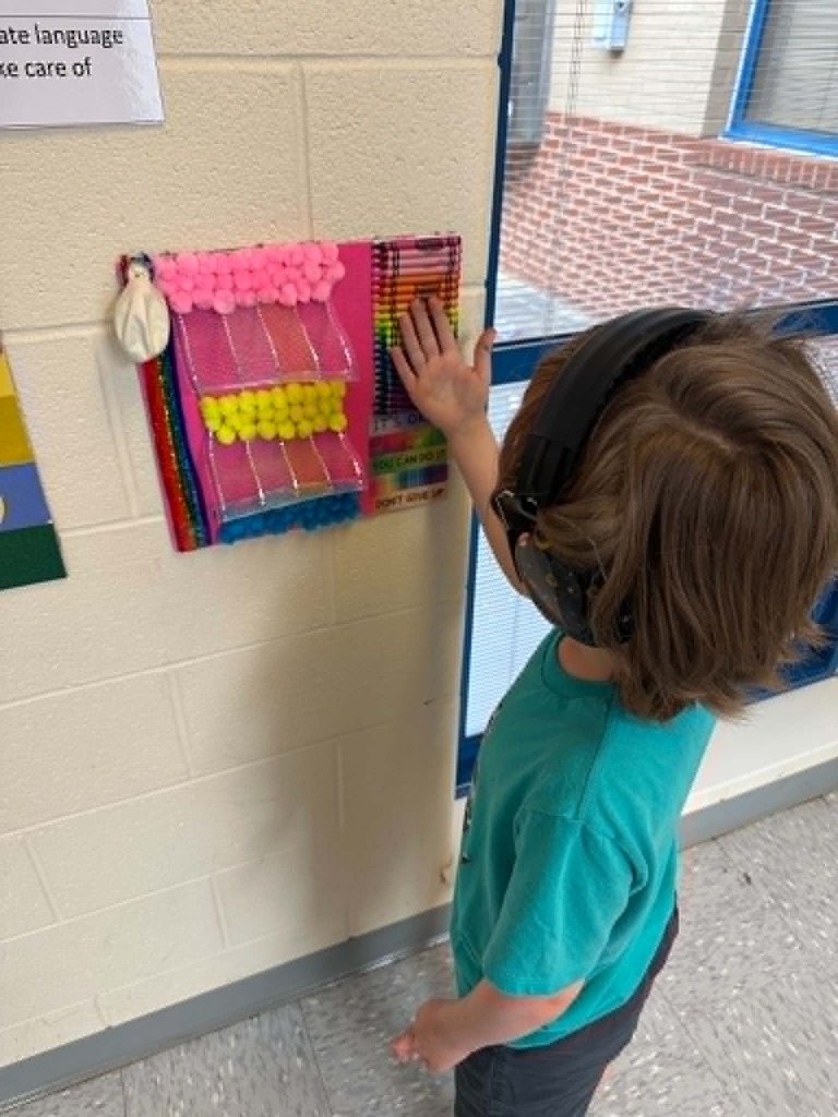 A child wearing headphones interacts with a tactile board featuring colorful materials on a classroom wall.