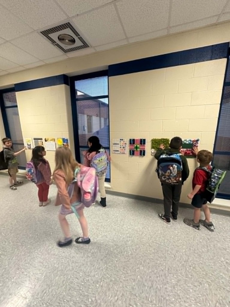 Several children with backpacks stand in a hallway, facing artwork on a wall with windows behind them.