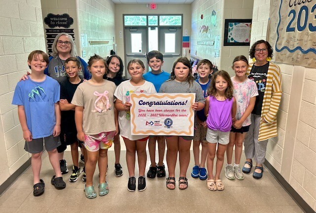 Nine children and three adults stand in a school corridor holding a sign with "Congratulations!" written on it.