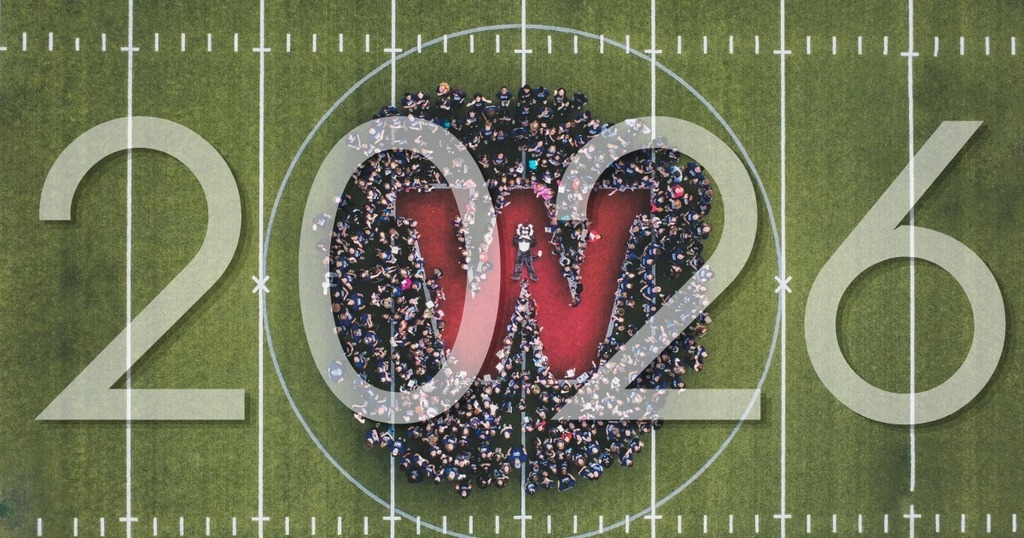 Overhead view of a football field with a heart shape formed by people, surrounded by "2026" in large numbers.