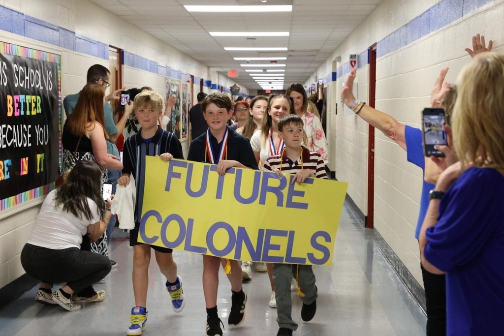 School hallway with children holding a yellow sign that reads "Future Colonels." People line the walls.