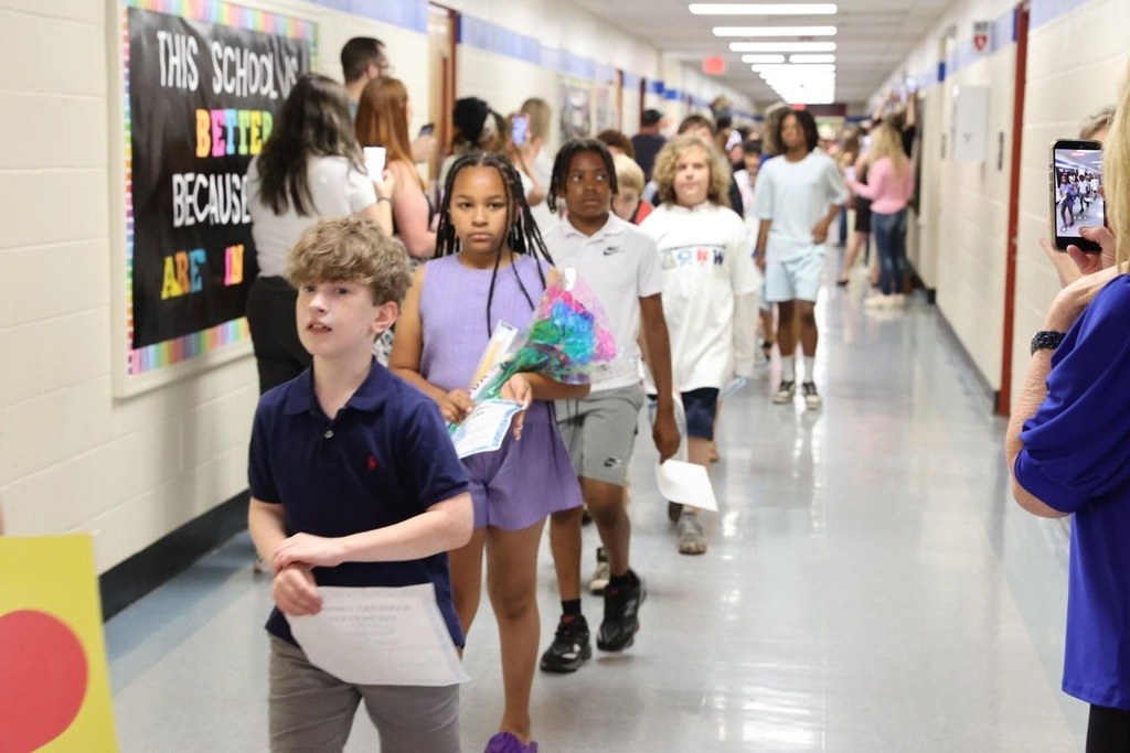 Children walk through a brightly lit school hallway, some holding papers. A person films with a phone.