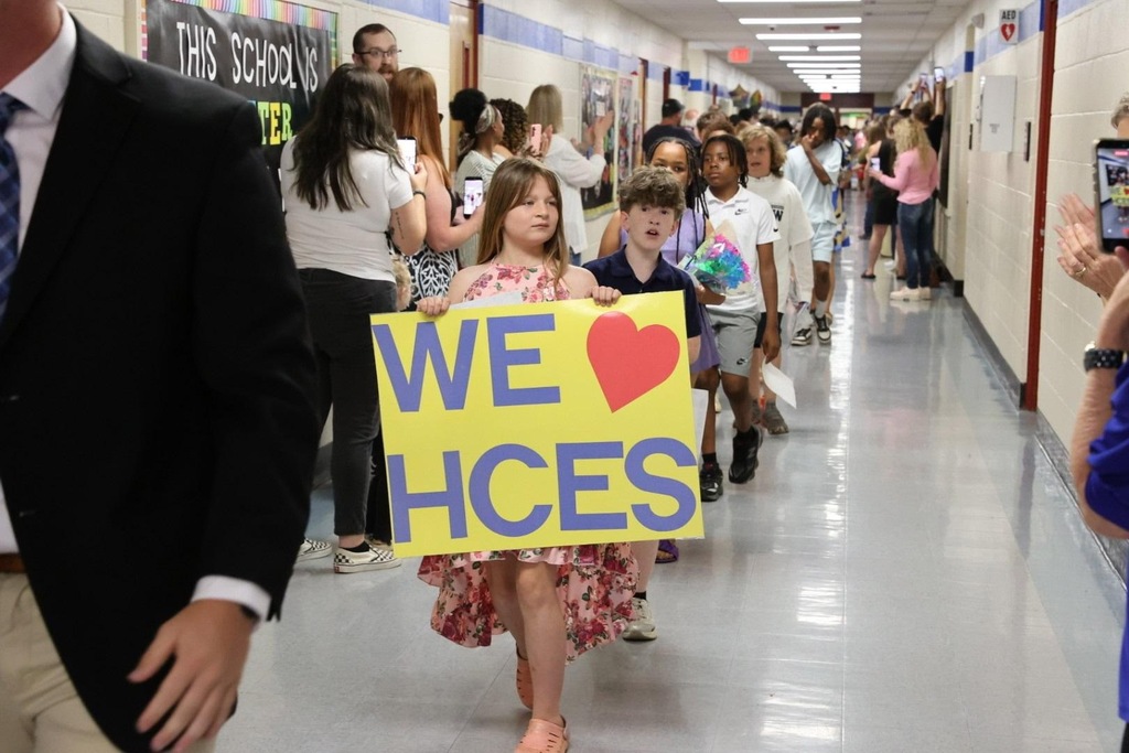 A group of students walks down a school hallway, with one holding a sign reading "WE ♥ HCES".