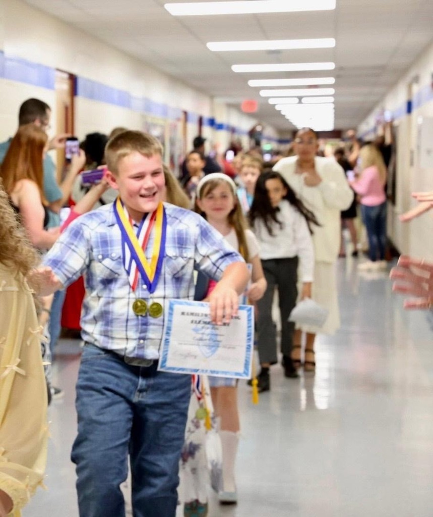 A young boy with a medal walks in a school hallway, holding a certificate. Several people are walking behind him.
