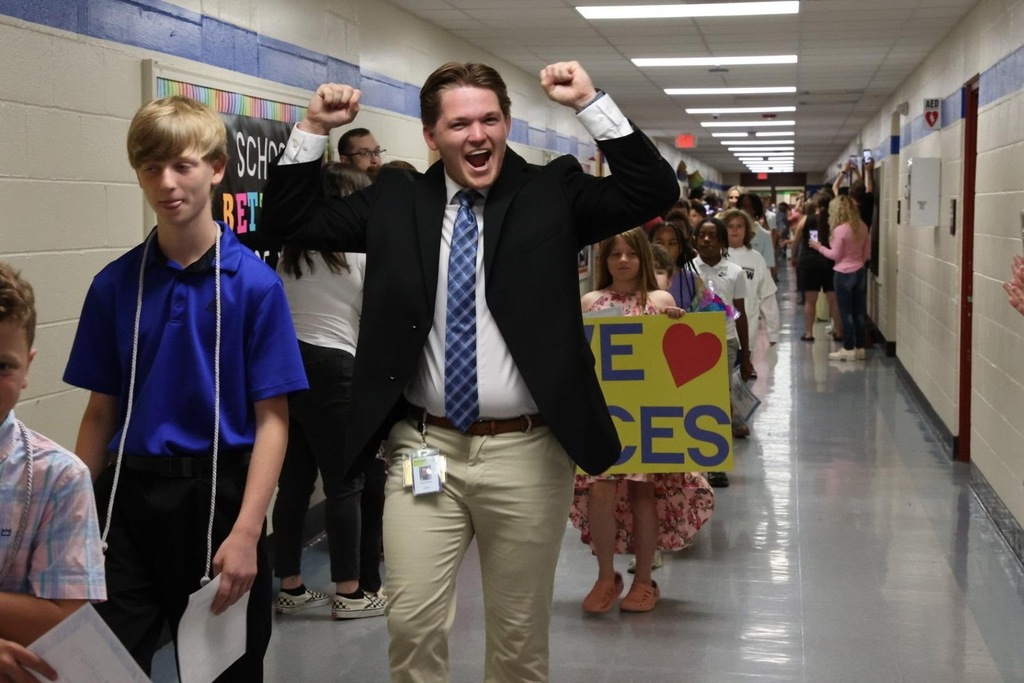 Man in a suit and tie cheering with others in a school hallway. Some hold signs.