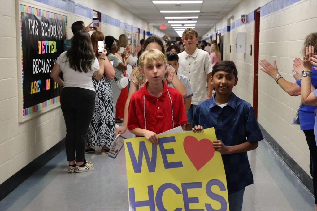 Two children hold a sign reading "WE HCES" in a school hallway. Other students and adults walk behind them.