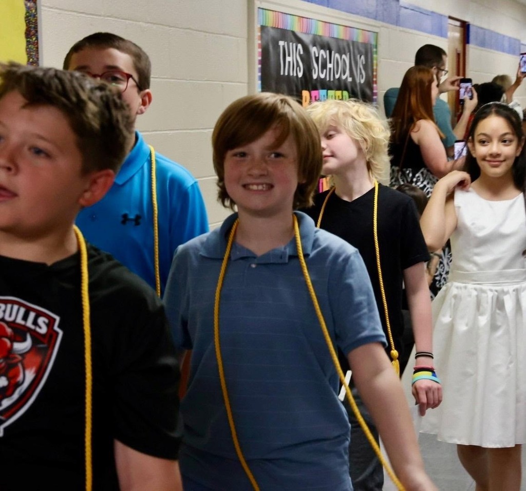 Several kids stand near a wall with a sign that reads "THIS SCHOOL". A boy wears a Bulls shirt, while others wear white and blue shirts.