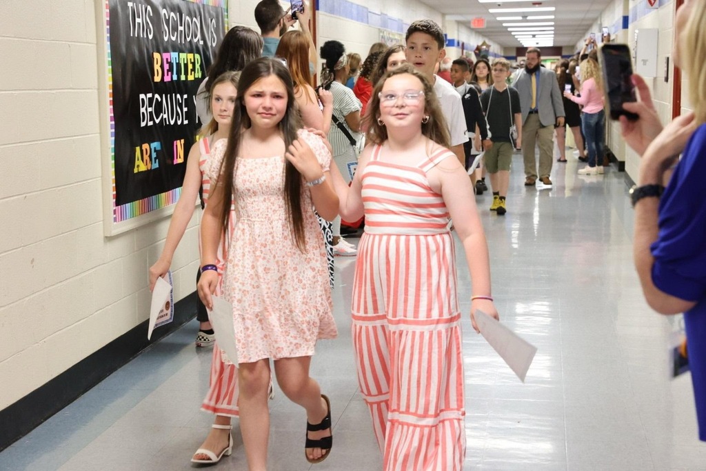 A school hallway with two girls in pink dresses walking and holding papers. Behind them, students are walking and a person is taking a photo.