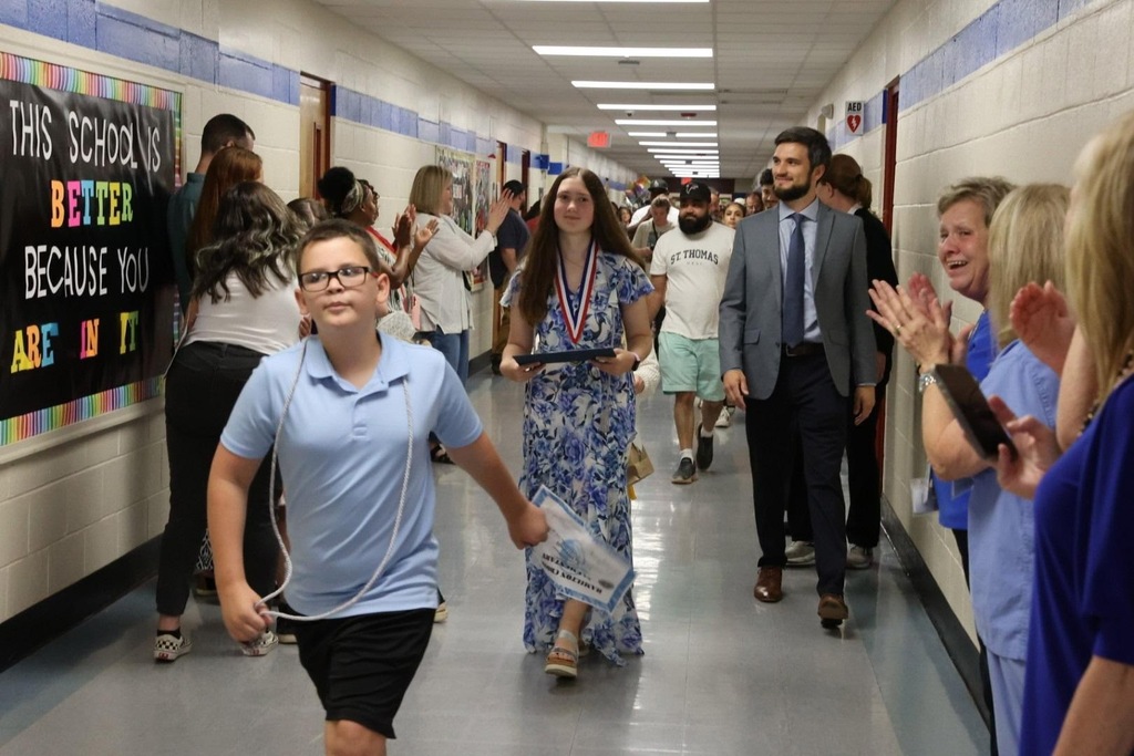Students and teachers walk in a hallway. A boy in a blue shirt holds a medal. Behind them, a bulletin board has colorful writing.