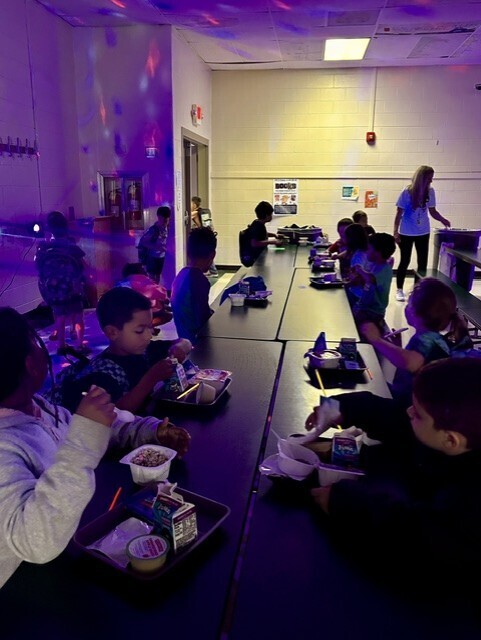 Children seated at tables in a room with purple lights and a woman in a white shirt standing.
