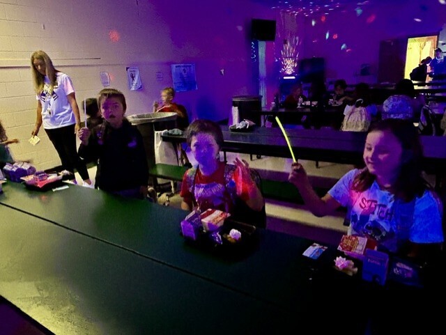 Three children sit at a table with food under a glowing purple light. A woman stands behind them.