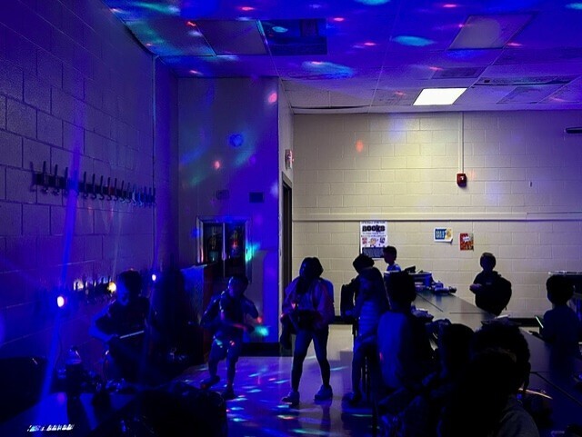 A room with purple lights on the ceiling and walls. People gather in the center with some standing and others seated.