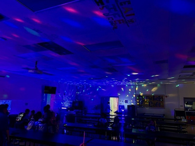 A room with purple lighting, tables and chairs. People are seated around tables, a fan is on the ceiling.