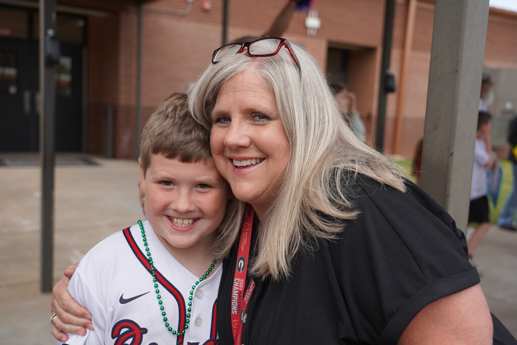 Woman and boy smiling. Boy wearing baseball team jersey and necklace. Background shows a building.