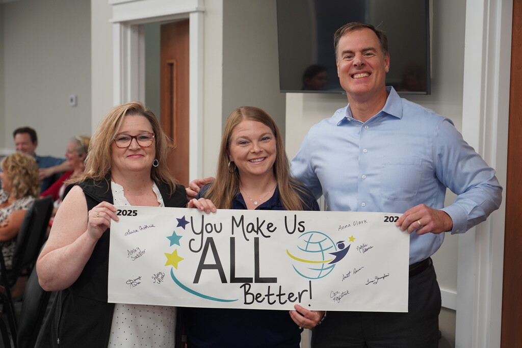 Three people pose for a photo with a sign. The sign reads, "You Make Us All Better!" The woman on the left wears glasses.