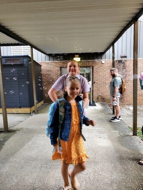 Two children, one with a backpack, walk on a covered walkway, with a person with a backpack standing behind.