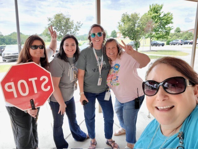 Five women standing, one holding a red stop sign, all wearing sunglasses. They pose for a photo with trees and parked cars in the background.