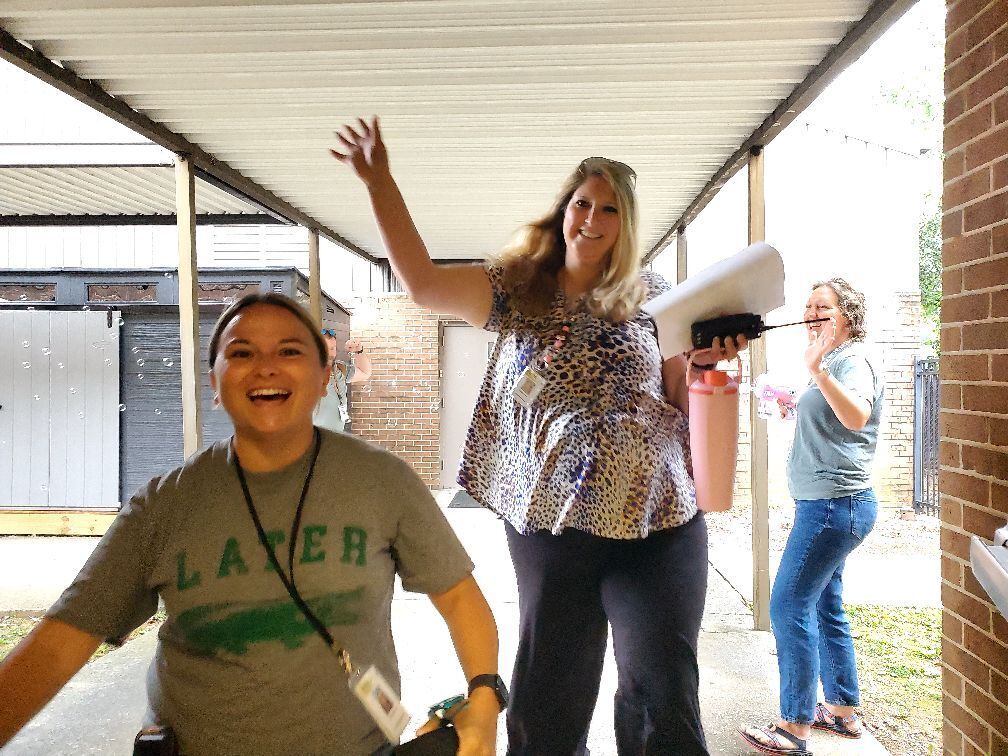 Three people in a hallway; one holds a paper roll, another laughs, and the third stands near a brick wall.