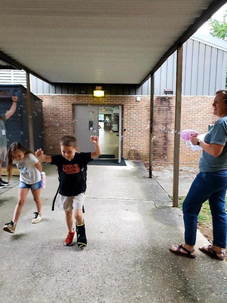 Three children under an awning. One child sprays bubbles, while the other two run. A woman stands nearby.