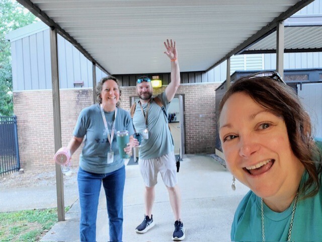 Three people smile and wave outside a building. Two hold drinks. The background shows a brick building and a fence.