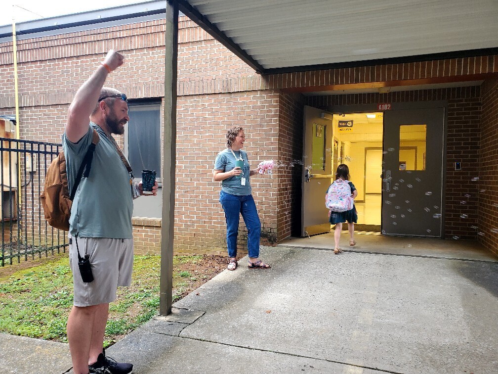 A man with a backpack waves at a woman and a child outside a school building.