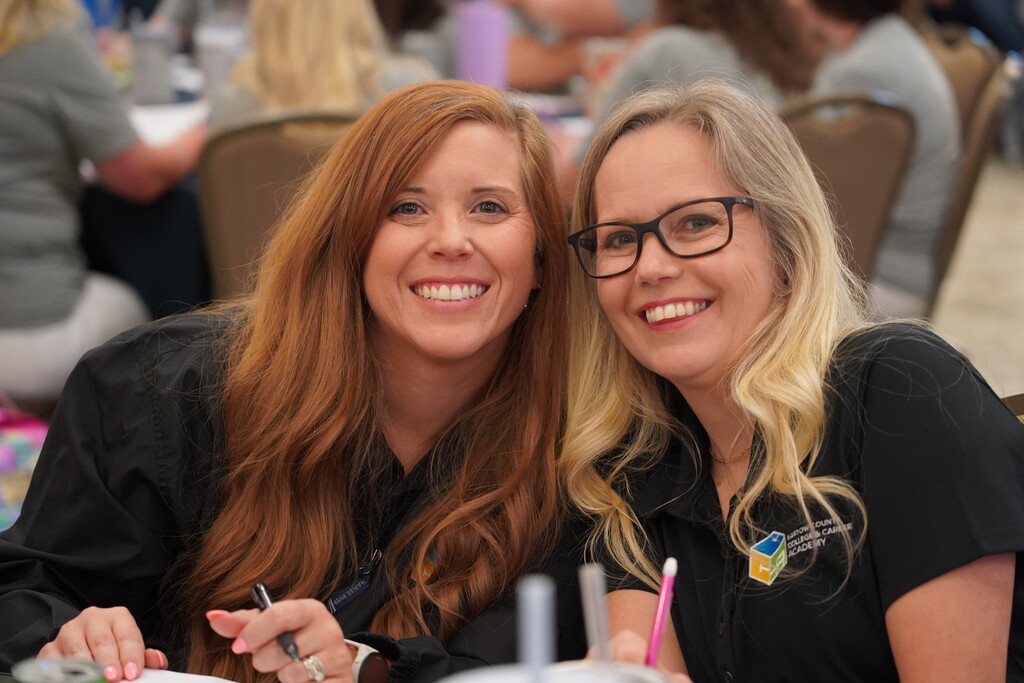Two women, one with long hair and the other with glasses, smiling and seated at a table.