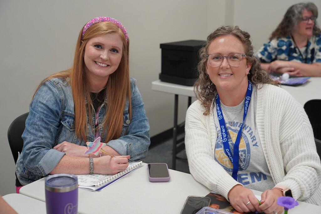 Two women sit at a table, one wearing a denim jacket and the other a white sweater, both smiling.