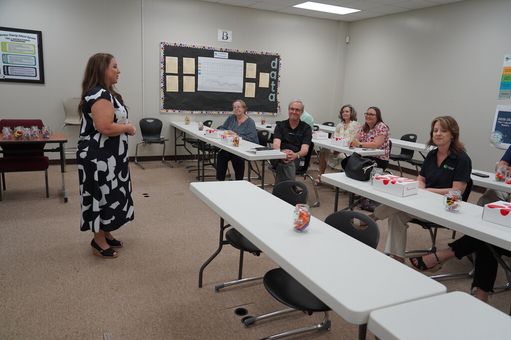 A woman stands in a classroom, addressing several seated adults at tables. A bulletin board and papers are in the background.
