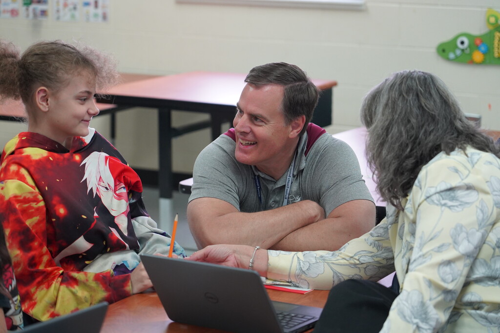 A man and two girls sit at a desk, smiling. One girl wears a colorful shirt, another a gray shirt.