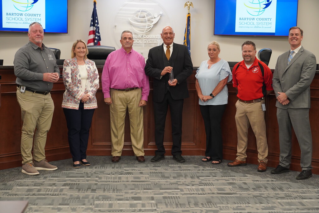 A group of seven people stands behind a desk, possibly at an award ceremony. A man in a red shirt holds an award.