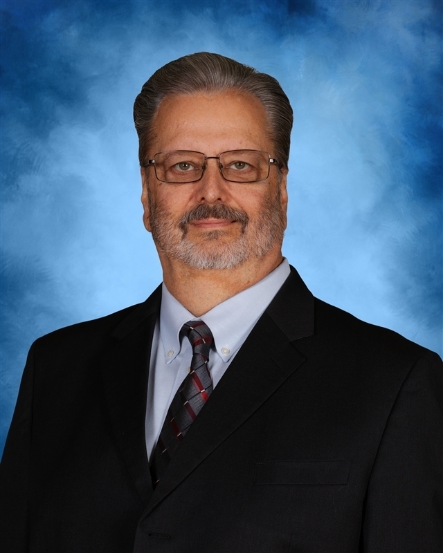 A man wearing a suit and tie with glasses and a beard stands against a blue background.