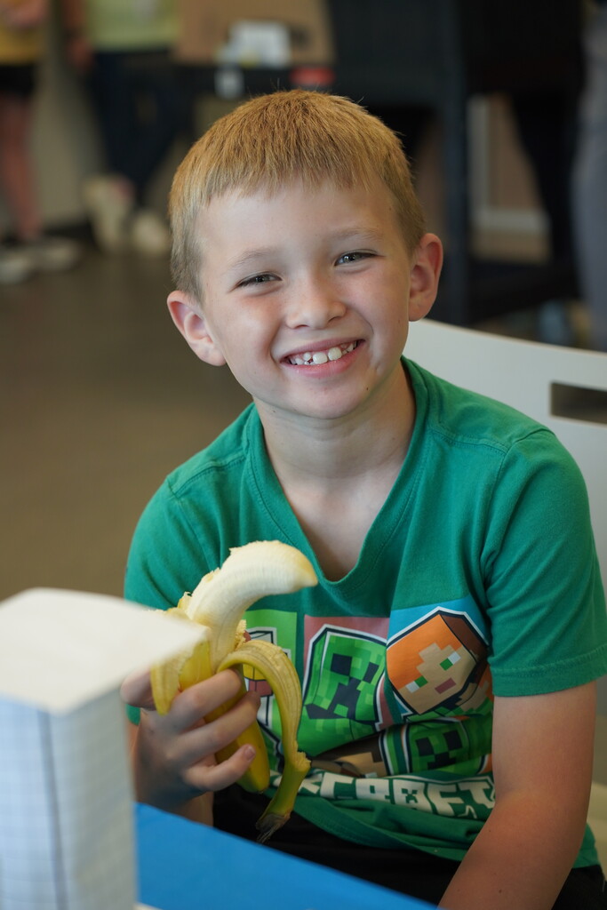 A young boy with blonde hair smiles and holds a banana in a classroom.