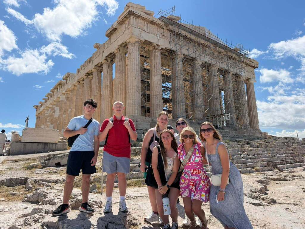 Six individuals in casual attire stand in front of the Parthenon, a classical Greek temple with columns, on a sunny day.