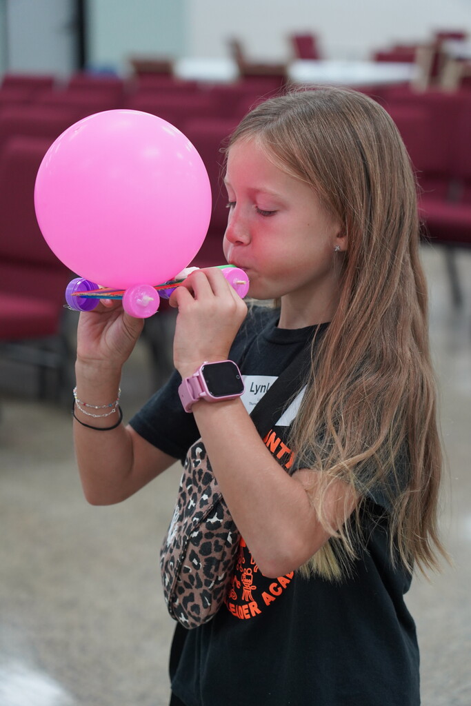 A girl blows up a pink balloon with a long nozzle, standing in a room with chairs.