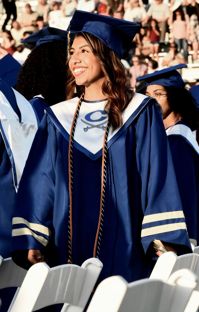 Woman in blue graduation gown with white stole, standing in row of chairs. Audience behind her is blurred.