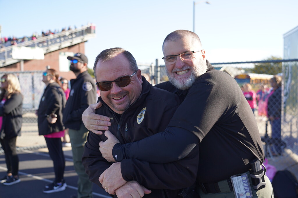 Two men hug each other at an outdoor event. They wear glasses and black clothing. A fence and people are in the background.