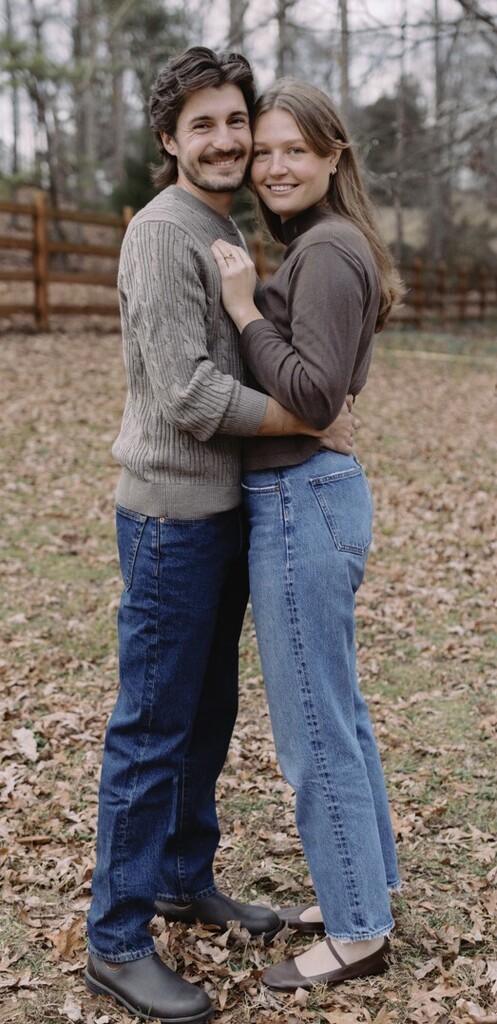 A man and woman stand close, smiling, in a yard with fallen leaves and a wooden fence.