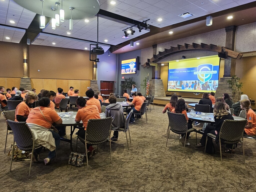 A conference room with large tables; people in orange shirts are seated, facing a large screen with a presentation.