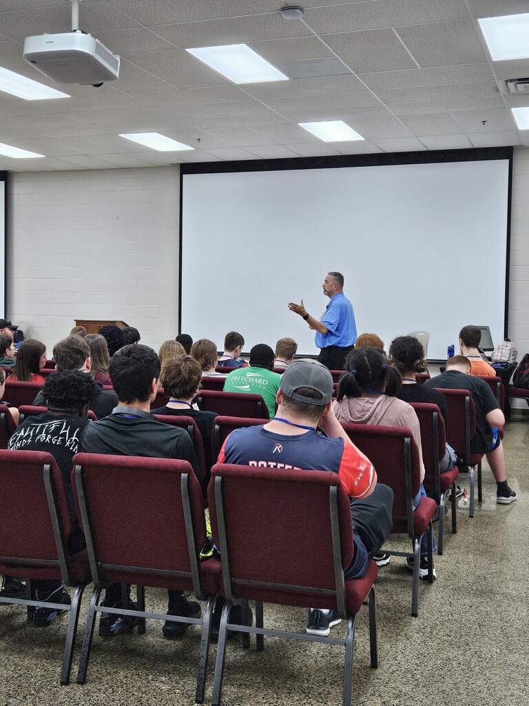 People seated in red chairs in a room with a projector screen, a man in blue stands in front of the screen.
