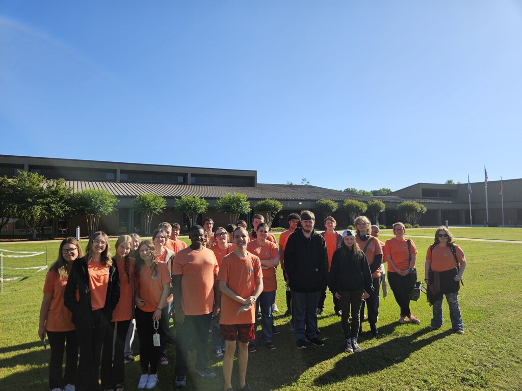 A group of people in orange shirts, with some wearing black, standing in a field with a building in the background.
