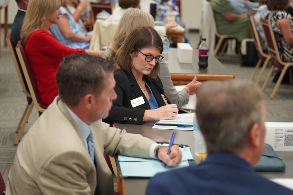 A group of people seated at a table during a meeting. A woman in a suit writes on paper.