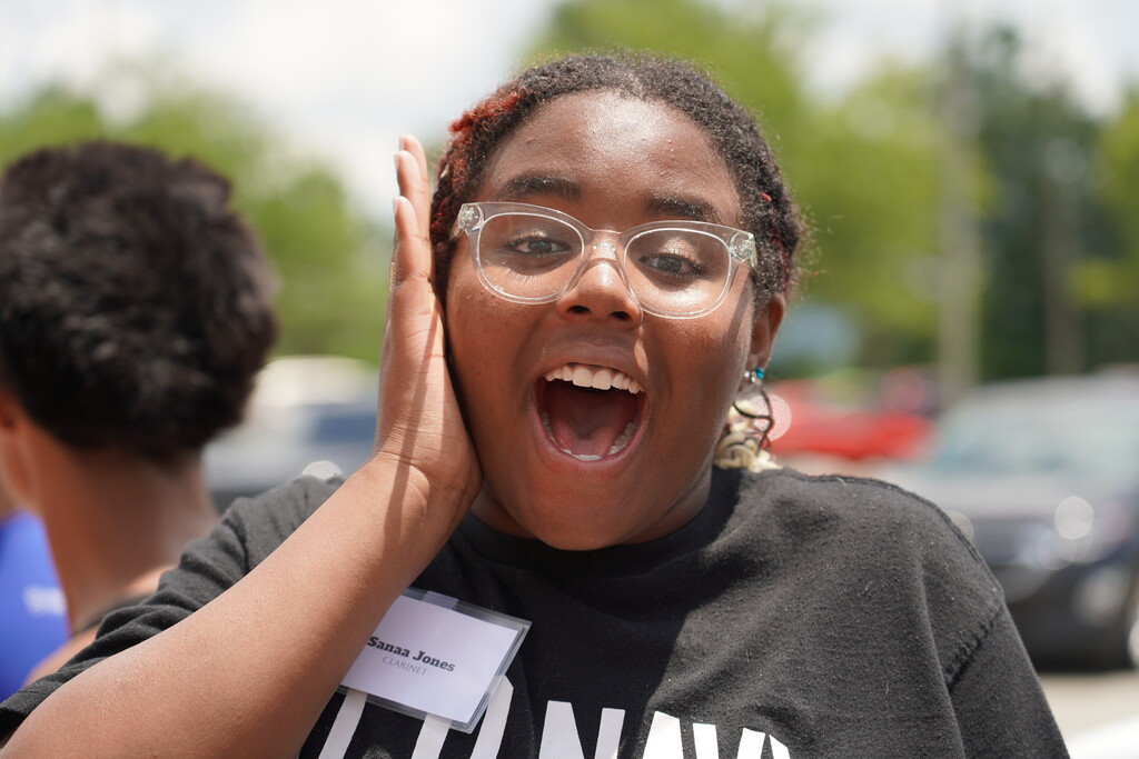 Woman in black t-shirt with white text and nametag, mouth open, holding hand to face, in a parking lot.